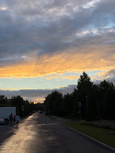 The image depicts a street scene in Zimyonki, Russia, at dusk. The sky is dominated by dramatic clouds, with a band of vibrant orange and yellow light from the setting sun illuminating the horizon. Dark, silhouetted trees line the right side of the street, which appears to be wet, reflecting the dim light.

In the foreground, a white van is parked on the left side of the road. Two people are visible near the van, with one standing and the other appearing to be walking away. Further down the street, other vehicles are parked and in motion, casting shadows on the wet asphalt. Streetlights are visible, along with a few traffic signs. The overall atmosphere is serene and somewhat melancholic, characteristic of the twilight hour. The weather appears to be calm, with the wet road suggesting recent rain.