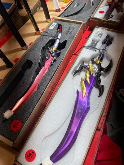 The image shows two prop swords laid out in cardboard boxes filled with foam packaging. Both swords are detailed with intricate designs. The sword on the left is predominantly red and black, with a curved blade that appears to be metallic. The sword on the right is purple and gold, with a more complex hilt and jagged blade design. Red circular stickers with white numbers and a euro symbol are visible on the foam packaging of both swords, indicating prices of "75€" and "79€". In the background, parts of other items are visible, including what appears to be furniture legs and boxes, suggesting a retail or display setting. The lighting suggests an indoor environment. The mention of Villepinte, France, places this scene within a specific geographic context, likely a convention, market, or retail store in that area.