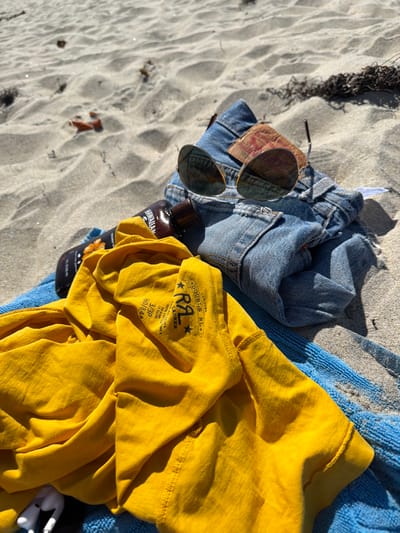 The image depicts personal items resting on a sandy beach under direct sunlight, presumably in Del Mar, United States of America.
In the foreground, a folded blue terrycloth towel is visible. On the towel, to the bottom left, a pair of white wireless earbuds are present. Over the towel, a crumpled yellow t-shirt is laid out; a label on the shirt clearly displays "LIMITED EDITION RRL DOUBLE RL RALPH LAUREN 3/P 160/84A". Partially obscured by the yellow shirt and resting on the towel is a brown plastic bottle with "HAWAIIAN" visible on its label, indicating a tanning or sun-care product. To the right of these items, a pair of blue denim shorts or jeans are folded, featuring a red fabric tab on the pocket. Resting on the denim are a pair of aviator-style sunglasses with gold frames and reflective lenses.
The background consists of light-colored sand with distinct ripples and a few scattered pieces of dark organic debris, likely dried seaweed. No individuals are present in the scene. The harsh shadows and bright illumination suggest clear weather conditions.