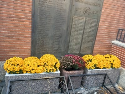 The image captures a solemn war memorial, likely situated in a public space, with a red brick wall serving as its backdrop. The central feature is a large, dark bronze plaque, titled prominently with the French phrase "MORTS POUR LA FRANCE" (Dead for France). Below this, the dates "1939" and "1945" are clearly visible, encompassing a circular emblem, indicating a World War II commemoration.
The plaque is inscribed with numerous names, arranged in columns, presumably listing those who perished. While many names are somewhat blurry, some in the left column are discernible, such as BOULANGER, CABOT, CLEMENT, COLLAT, DUMONT, GASTON, LAFONT, LECLERC, MARTIN, MOREL, PETIT, PHILIPPE, RICHARD, and TALON. The right column also contains lists of names.
At the base of the memorial, several large planters hold an abundance of chrysanthemums. Three granite planters on the left are filled with vibrant yellow chrysanthemums, creating a bright contrast against the dark memorial and brickwork. To their right, a single, darker-toned pot holds chrysanthemums of a deep purple or burgundy hue. In the immediate foreground, two decorative metal grates with swirling patterns are placed, one partially covering the ground in front of the planters.
The overall scene conveys a sense of remembrance and respect, typical of war memorials in France, especially with chrysanthemums often associated with All Saints' Day. The lighting suggests it is daytime, possibly under an overcast sky.