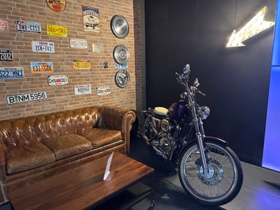 The image depicts an interior space in Alcobendas, Spain, styled with an automotive and motorcycle theme. On the left, a tufted brown leather Chesterfield-style sofa occupies the foreground, facing a rectangular wooden coffee table with a small white informational stand. The wall behind the sofa is exposed brick, adorned with approximately twenty-three assorted international license plates, including examples from Texas, Nevada, Utah, Wisconsin, and China, along with several metallic circular hubcap-like decorations and a "Mustang" vintage-style tin sign. To the right, a purple and chrome cruiser-style motorcycle, consistent with a Harley-Davidson aesthetic, is prominently displayed. The motorcycle features spoked wheels, a single headlight, and a white or light-colored seat. Behind the motorcycle, the wall is dark and textured, featuring a large, yellow illuminated arrow sign pointing downwards and to the left. The lighting is artificial, illuminating the various decor elements and the motorcycle. No individuals are present in the scene.
