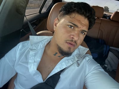 A close-up, front-facing selfie shows a man with dark, wavy hair and a goatee, wearing an unbuttoned white dress shirt. He has a tattoo on his neck and appears to be in the passenger seat of a car. The interior of the car features brown leather seats, and a seatbelt is visible. The background shows a window with a blurred view of trees and a road, suggesting he is driving through a rural area. The image is a portrait of the man, focusing on his face and upper body. The provided location details about Shtarkovo, Bulgaria, are not visually inferable from the image itself.