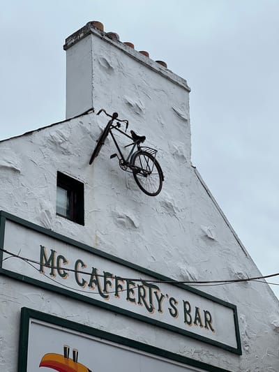A vintage bicycle is mounted on the exterior wall of McCafferty's Bar in the town of Donegal, Ireland. The bicycle, which appears to be dark in color, is positioned on the steeply sloped roofline of the white stucco building. One pedal is extended, as if in motion, and the bike's handlebars are angled upwards. Below the bicycle, a sign for "McCafferty's Bar" is visible, with green and orange lettering. The building has a rough, textured finish and a small, dark-framed window is visible to the left of the bicycle. The sky above is overcast and grey.