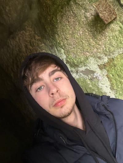 The image displays a head-and-shoulders selfie of a young adult male. He has light brown hair, green eyes, and light stubble on his chin and upper lip. He is wearing a dark-colored hooded sweatshirt with the hood pulled over his head, underneath a dark blue or black puffer-style jacket. The subject looks directly into the camera with a neutral expression. The background consists primarily of a rough, textured rock surface, extensively covered in green moss and lichen. In the upper right quadrant of the background, a small, roughly square, dark brown object, possibly a weathered piece of metal or a distinct rock discoloration, is visible against the mossy rock. The left side of the image background is significantly darker, indicating deep shadow or an enclosed space. The specific location within the city of Ribeira de Pena, Portugal, cannot be determined from the visual evidence provided.