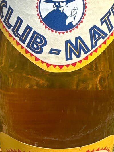 The image shows the top portion of a glass bottle with a yellow and red label reading "CLUB-MAT". The label includes a circular emblem with a depiction of a person in a dark blue robe and a wide-brimmed hat, holding a glass of liquid. The bottle is filled with a light brown liquid, likely a beverage, and has a translucent appearance. The image is cropped, only showing the label and a small portion of the bottle's contents. There are no people or other objects visible in the image besides the bottle itself. The coordinates 50.847, 4.383 place the location in the center of Brussels, Belgium, but there is no information available on the location within the city.