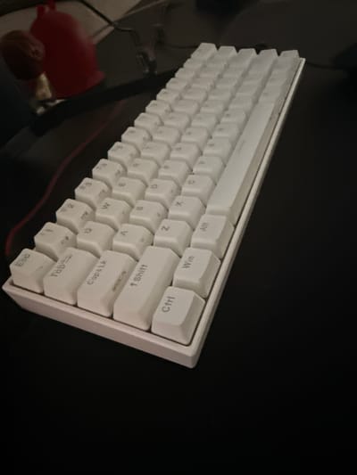This is a close-up, angled shot of a white mechanical keyboard on a dark surface, likely a desk. The keyboard is oriented diagonally across the frame, with the top left corner closest to the viewer. The keys are white and appear to be made of plastic. Some keys have visible legends. From top to bottom, and left to right, some of the visible key legends include: "Win", "Ctrl", "Shift", "CapsLk", "Tab", and "Esc". The keyboard has a minimalist design, with a thick bezel. The lighting appears to be artificial, casting some shadows and highlighting the texture of the keys. In the background, to the left of the keyboard, there are some blurry objects, including a red item and a brown, nut-like object. The overall scene suggests a personal workspace, possibly at home or in an office. There are no people visible. The time of day and weather are not discernible from the image, and there are no specific location cues to indicate it is in Vespolate, Italy, other than the provided context.