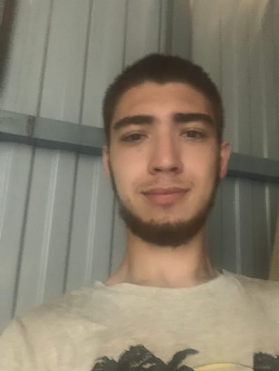 This media file is a portrait of a young man with short, dark hair and a beard. He is looking at the camera, with a neutral expression. He is wearing a light-colored t-shirt. The background appears to be an indoor setting with corrugated metal walls, possibly a garage or shed. The lighting suggests it is daytime. There is no visible text in the image.