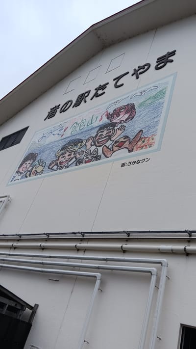 A mural depicting cartoon characters is painted on the white exterior wall of a building. The mural is located in Tateyama, Japan, as indicated by the Japanese text at the top of the mural, which reads "館山の駅たてやま". The mural features several figures with smiling faces and exaggerated features, some appearing to be children and others more fantastical creatures. The background of the mural is a wavy blue pattern, suggesting water or sky, with some red and orange elements on the right. Below the main image, a smaller line of Japanese text reads "画: さかなクン", which translates to "Drawing: Sakana-kun", likely crediting the artist. The building's wall is textured, and pipes are visible running horizontally along the lower section of the exterior. The sky above is overcast.
