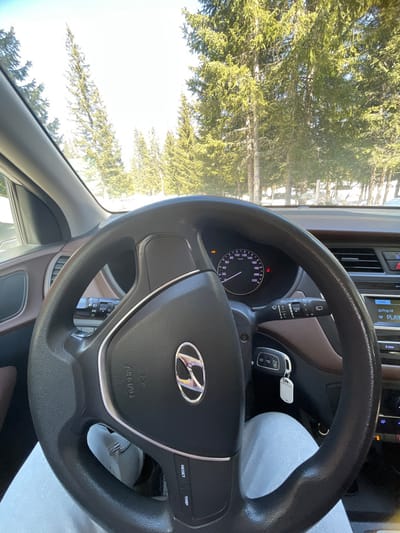 The image depicts the interior of a Hyundai vehicle from the driver's perspective, stationary in Palež, Montenegro. The driver's legs, clad in light grey sweatpants, are visible below the black multi-function steering wheel, which features a silver Hyundai logo and "AIRBAG" inscription. The instrument cluster, showing a speedometer at 0 km/h and illuminated warning lights including a yellow engine check light, is visible through the steering wheel. A set of Hyundai car keys with a white fob is inserted into the ignition. To the right, a portion of the car's infotainment system displays "Pop M" and "PLAY". The dashboard is a combination of brown and black materials. Through the windshield, a sunny outdoor winter scene is visible, dominated by numerous tall green coniferous trees, with snow covering the ground. A light-colored wooden fence or barrier is partially discernible among the trees on the right side of the background.