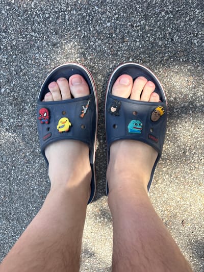 A top-down, eye-level medium shot captures a person's feet wearing dark blue Crocs on a concrete surface. Each shoe is adorned with several colorful Jibbitz charms. The left shoe features a Spider-Man charm, a yellow duck charm, and an assault rifle charm. The right shoe displays a Batman charm, a Pepe the Frog charm, and a charm depicting a person wearing a crown. The concrete ground appears to be asphalt with small grey and white pebbles. Sunlight casts shadows on the ground, suggesting an outdoor setting. The image does not provide any information to locate it in Aeroflotsky, Ukraine.