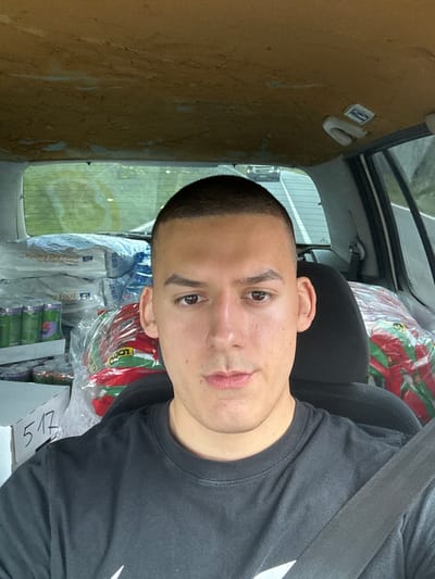 A close-up, candid photograph depicts a man wearing a black t-shirt with a seatbelt across his chest. He is situated in the passenger seat of a vehicle filled with various goods, possibly pet food and packaged items. The interior of the vehicle is visible, with a worn, possibly older interior finish. Outside the window, a blurred urban scene is discernible. There's no verifiable evidence in the image that confirms it's taken in Medovo, Bulgaria.