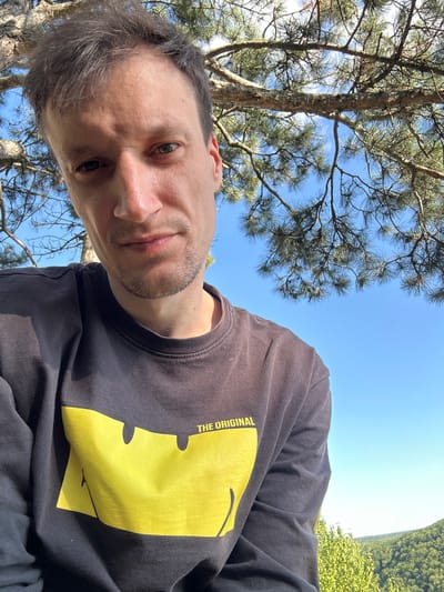 A man is shown in a selfie from the chest up, with trees and a clear blue sky in the background. He is wearing a dark long-sleeved shirt with a large yellow graphic on the front, which appears to be a stylized smiley face. The text "THE ORIGINAL" is visible above the graphic. The man has short brown hair and stubble. The lighting suggests it is daytime and sunny. The scene is outdoors, possibly in a park or a wooded area. There is no identifiable location in the city of Nihyn, Ukraine mentioned or visible in the image.