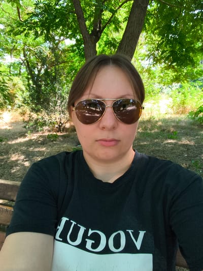 A medium shot is taken of a young adult with light brown hair, wearing large, gold-rimmed aviator sunglasses and a black t-shirt with white lettering. The person is seated on a wooden bench in an outdoor setting with lush green trees and foliage in the background. The sun is casting dappled shadows on the ground. The overall impression is of a casual moment captured outdoors. The location is identified as Bagerovo, Ukraine.