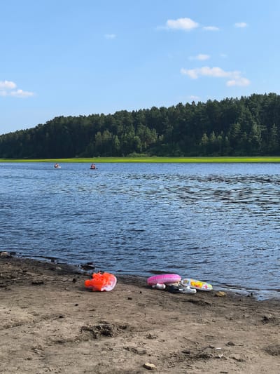 In Sysert, Russia, on a sandy shore adjacent to a wide body of water, an orange inflatable float and a pink inflatable ring lie on the ground. Nearby, two white Crocs and a yellow and green inflatable float are also visible on the sand. The water is calm with ripples and reflects the blue sky, which is dotted with a few white clouds. In the distance, a dense green forest borders the far side of the water, with a strip of bright green vegetation separating the trees from the water's edge. Two individuals are present in the water, each on an inflatable mat, further out from the shore.