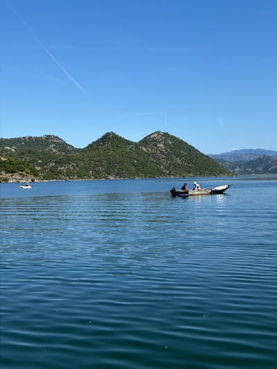 This image depicts a serene daytime scene over a large body of water, likely a lake, in the vicinity of Kruševica, Montenegro. The sky is a clear, bright blue with a single, prominent white contrail traversing it diagonally from the upper left. In the background, verdant, tree-covered hills rise from the water's edge, exhibiting rugged, rocky outcrops. Behind these immediate hills, further mountain ranges with lighter, hazy blue tones are visible on the horizon, suggesting distance.
The water, a deep blue-green, is calm with numerous small, parallel ripples stretching across its surface, reflecting the sunlight. Two boats are present on the water. To the left, a solo individual is seated in a small, white boat, resembling a kayak or canoe, oriented towards the right. This person appears to be wearing a light-colored hat and top.
Further to the right and closer to the center of the frame, a larger, traditional-style boat is stationary. This boat is light-colored, possibly white or grey, with an outboard motor mounted at its stern. Two individuals are inside this boat. The person on the left is wearing a dark cap and a dark shirt. The person on the right is wearing a white cap and a white shirt, positioned towards the front of the boat, seemingly interacting with something on board. The boat appears to be well-used, showing some wear and tear on its hull. The overall impression is one of peaceful outdoor activity, possibly fishing or recreation, amidst a natural landscape.