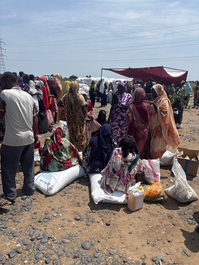 The image depicts a scene of people gathered outdoors, possibly receiving aid or provisions. The subjects are a mix of individuals, many of whom are wearing colorful clothing and headscarves. They are seated or kneeling on a ground covered with pebbles and dirt.
Several large sacks, likely containing food items like flour, are visible, along with a plastic jug of cooking oil and other supplies in a cardboard box. Some people appear to be tending to or sorting through these items. The presence of the sacks and the organized arrangement of goods suggest a distribution or collection point for humanitarian aid.
In the background, a large, dark red canopy provides shade, under which more people are gathered. The sky is visible and appears partly cloudy, suggesting daylight, likely midday given the lighting and shadows. Power lines stretch across the sky, indicating proximity to some infrastructure. The overall atmosphere seems somber and focused on the distribution of resources.
Visible text on one of the sacks reads "طحين FLOUR" and "NOT FOR SALE". Another piece of text on a box mentions "Refined Palm Olein" and "Country of Origin: Sudan". These details clearly identify the location and the nature of the distributed goods.