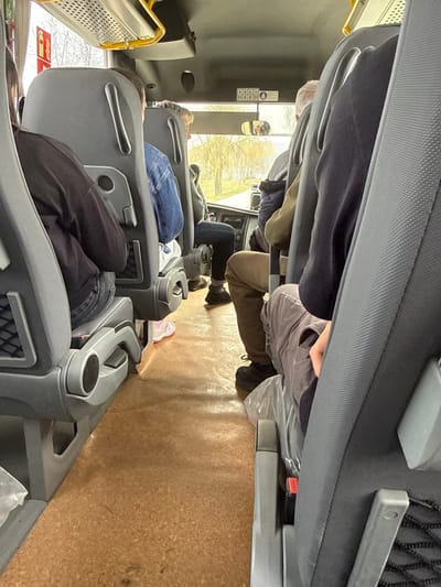 The image depicts the interior of a bus or minibus traveling in Dalbok Dol, Bulgaria, viewed from a rear-right passenger seat looking forward. Six individuals are present: four passengers are seated ahead of the vantage point, one is in the foreground right, and one is the unseen photographer.
On the immediate left, a passenger with dark hair wears a dark long-sleeved top and blue jeans, seated in a grey upholstered seat with a visible seatbelt buckle. A white fire extinguisher is mounted on the wall above this seat. Further forward on the left, a person with light brown hair wears a blue denim jacket and blue jeans, seated. A pink shoe is visible on the light brown, textured floor between the seats.
On the right side, a passenger wearing light brown trousers and dark shoes is seated directly in front of the photographer. A transparent plastic bag is on the floor near their feet. Further forward on the right, a person with grey hair is partially visible, seated in a similar grey seat.
The vehicle's interior features a light brown cork-like floor covering, grey upholstered seats with integrated cup holders and mesh pockets, and grey plastic seat frames. Overhead, a white ceiling with ventilation grilles and yellow handrails is visible. A small white sign with black pictograms, including a "no standing" symbol and a seatbelt symbol, is positioned above the windshield. Through the front windshield, a road is visible, flanked by trees with light green or yellow foliage, suggesting a natural outdoor environment. A rearview mirror reflects a blurred interior view. No explicit interactions between individuals are visible; all appear to be seated and facing forward.