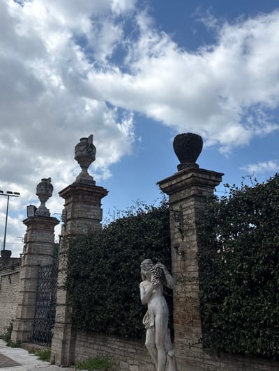 The image depicts an exterior scene in Foligno, Italy, under a partially cloudy blue sky. In the foreground, positioned right of center, stands a weathered light-colored stone statue of a partially draped female figure holding a cluster of grapes near her left ear. Behind and to the left of the statue, a tall, dense green hedgerow forms a property boundary, supported by a low brick and stone wall.
Three substantial brick and stone pillars are visible from left to right. The leftmost pillar, partially obscured, is topped with a grey stone urn and features a light fixture. A section of a dark, ornate metal gate is visible between this pillar and the central pillar. The central pillar is also topped with a grey stone urn, featuring decorative carvings. The rightmost pillar, positioned behind the statue, is topped with a dark, ovoid-shaped topiary and has a black, scroll-work metal fixture mounted on its side. A light pole with a black rectangular fixture is present in the far left background. The ground in the lower-left foreground consists of a paved surface with sparse weeds growing along its edge.