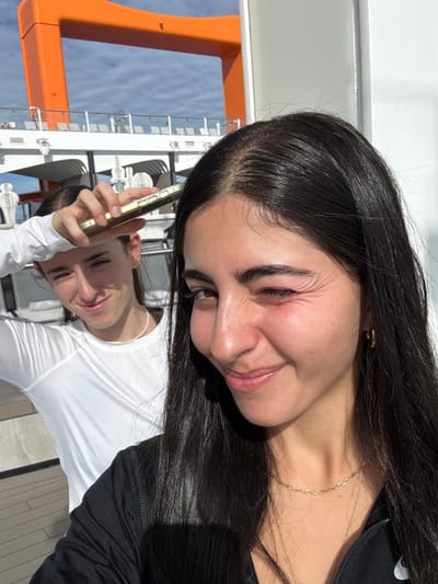 Two young females are depicted on the outdoor deck of a large vessel, consistent with a cruise ship, located near Long Island, Bahamas. The subject in the foreground, with long, dark hair, wears a black top, a thin gold chain necklace, and a small gold hoop earring on her visible left ear. She is winking her right eye, showing prominent periorbital wrinkles, and smiling towards the viewer. Her facial skin exhibits a reddish tone. Behind and slightly to her left, a second female subject with dark hair, pulled back from her face, wears a white long-sleeved athletic shirt and a white beaded necklace. She is also smiling and is seen holding a light-colored smartphone horizontally above her forehead, using it to shade her eyes. The background reveals the white superstructure of the vessel, including railings of an upper deck, and a distinctive orange, arch-like structural component against a partly cloudy blue sky. The scene is brightly illuminated by natural daylight.
