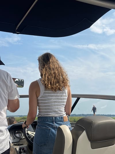 A photograph captures a scene from a motorboat, positioned in the city of Montgomery, United States, likely on a body of water such as a river or large lake. The primary subject, a female with wavy light brown hair, is seen from behind, wearing a horizontally striped white, black, and yellow tank top and a blue denim skirt. Her left hand rests on the boat's black steering wheel. A dark-colored smartwatch is visible on her left wrist. To her left, the partial back of a male in a white t-shirt is visible. A rectangular side-view mirror, affixed to the boat, reflects a distant, multi-story classical-style building with columns, flanked by trees. In the far background, beneath a clear blue sky dotted with sparse white clouds, a solitary male figure wearing a light hat and dark clothing walks on a low-lying, vegetated landmass. The interior of the boat shows a beige and grey upholstered seat in the foreground and a dark blue canopy overhead. The boat appears to be stationary or moving slowly on calm water.