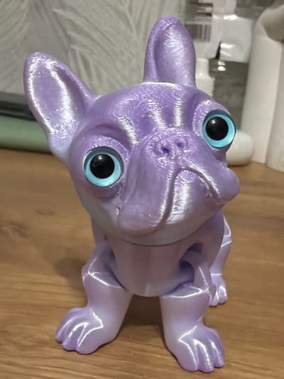 A close-up, top-down shot reveals a light purple, 3D-printed figure of a dog, possibly a French Bulldog, with large, bright blue eyes. The dog is lying on a wooden surface. The background is blurred but appears to show household items, including a white container and a tiled surface. There's no discernible information to suggest that this photo was taken in Staraya, Russia.
