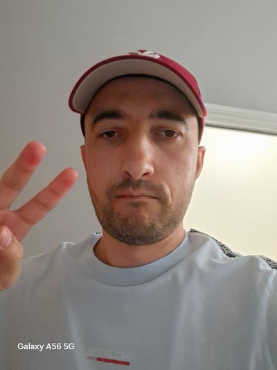 A medium shot captures a man wearing a red and white baseball cap and a light blue t-shirt. He has a light complexion, brown eyes, and a short, stubbly beard. His left hand is raised, with his index and middle fingers extended in a peace sign. The man appears to be indoors, with a plain, light-colored wall behind him. The bottom right corner of the image displays text that reads "Galaxy A56 5G". The context provided does not suggest this image is related to the city of Greater London, United Kingdom.