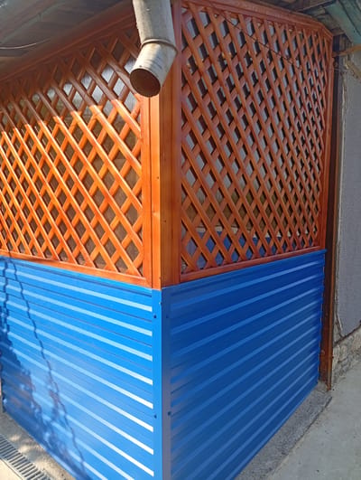 This image captures a close-up view of a corner of a building's exterior, likely a gazebo or an extension, constructed with two distinct materials. The lower portion is clad in bright blue corrugated metal siding, reflecting sunlight and casting subtle shadows. This siding appears to be anchored with visible screws. Above this blue base, a lattice-like wooden structure with a warm, varnished finish forms the upper section. The lattice is arranged in a diamond pattern, and behind it, a translucent material, possibly polycarbonate sheeting, is visible, allowing some light to filter through. A weathered metal downspout is attached to the wooden lattice structure and extends downwards. The overall scene suggests an outdoor setting, possibly a backyard or a utilitarian area, given the presence of the drain grate in the lower left corner. The lighting indicates it is daytime, with bright sunlight illuminating the scene. There is no text visible in the image. The construction materials and style are consistent with the rural or suburban architecture found in locations like Podvirne, Ukraine.