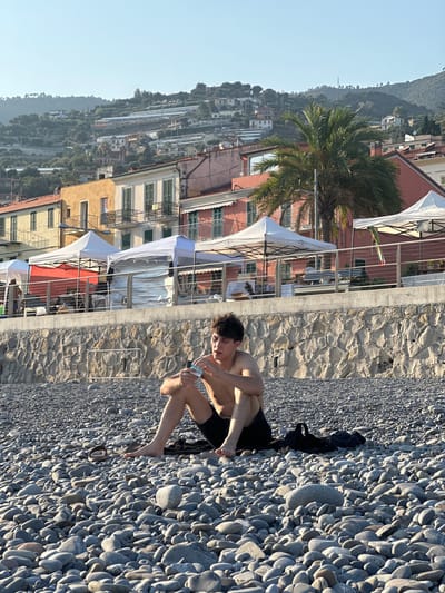 The image shows a young man sitting on a pebble beach in Ospedaletti, Italy. He is wearing black swim trunks and is shirtless, holding a plastic bottle of water. In the background, there are buildings on a hillside, some with colorful facades, and a palm tree. There are also several white and red tents set up along the promenade. The sky is clear and blue. The beach is covered in smooth, grey pebbles.