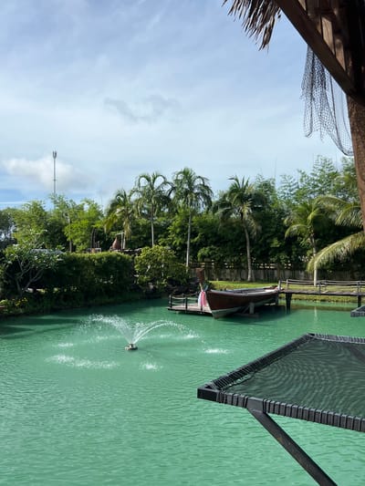 The image captures a tranquil scene in Tambon Pa Kae Bo Hin, Thailand, likely during the daytime. The foreground is dominated by a wooden structure with a dark net, suggesting it might be part of a canopy or a shaded area, with some palm fronds visible on the left.
The main focus is a body of turquoise water, possibly a pond or a canal, with gentle ripples on its surface. A small, automated fountain is active in the water, creating a spray of white water that contrasts with the green hue of the water. Along the edge of the water, lush green vegetation and trees, including several palm trees, create a dense, tropical backdrop.
A wooden pier extends into the water, and a traditional wooden boat is moored alongside it. The boat appears to be empty, adding to the peaceful ambiance. Further in the background, a tall telecommunications tower is visible against a partly cloudy sky, indicating a blend of natural and modern elements in the landscape. The overall mood is serene and relaxed, embodying the peaceful atmosphere of a tropical setting. No visible text is present in the image.