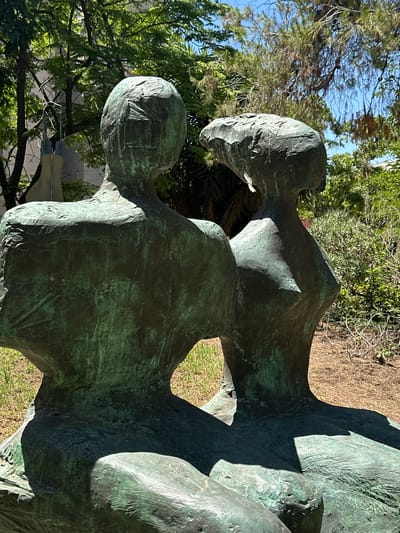 The image depicts a bronze sculpture of two figures, appearing to be seated and positioned in close proximity to one another, with their backs largely to the viewer. The figures are abstract and stylized, with smooth, rounded forms characteristic of modern sculpture. Their heads are distinct, one with a more rounded, cap-like shape and the other with a more elongated, somewhat flattened top. The surface of the sculpture exhibits a patina, with areas of green oxidation typical of aged bronze. The sculpture is situated outdoors, with dappled sunlight illuminating its surfaces and casting shadows. In the background, lush green foliage and trees suggest a park or garden setting. The ground around the sculpture is covered with dry grass and some sparse vegetation. The overall scene is tranquil and highlights the artistic form against a natural backdrop. The location is the city of al-Eizariya, Palestinian Territories.