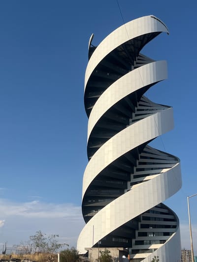 The image shows an architectural structure in Tlaxcalancingo, Mexico. It is composed of a series of large, white, curved bands that spiral upwards around a building with black and grey vertical supports. The bands appear to be constructed from individual, rectangular tiles. The sky is visible in the lower half of the frame, transitioning from a clear, deep blue to a paler blue with some clouds near the horizon. To the right of the building, there is some ground cover with sparse vegetation and a small tree. The photo is taken from a low angle, emphasizing the height and scale of the structure.