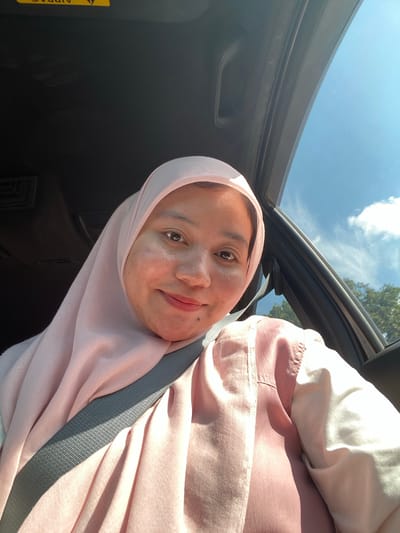 A woman wearing a light pink hijab and a pink top is seated inside a car, with the camera positioned to capture a selfie. The interior of the car is dark, but the bright blue sky with scattered clouds is visible through the open window on the right. Trees can be seen in the background outside the car. A grey seatbelt crosses diagonally over the woman's chest. The scene appears to be in Mandi Angin Barat, Indonesia, evidenced by the visible trees and sky characteristic of the region.