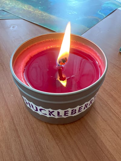 A lit, single-wick candle contained within a cylindrical, grey metal tin with a silver rim is positioned on a light brown wooden surface. The candle features deep reddish-pink wax, with a prominent circular pool of melted wax surrounding the actively burning wick. The flame is elongated, yellow-orange, and casts a bright glow, with its reflection visible in the melted wax. A white, irregularly shaped label on the exterior of the tin displays the partial word "HUCKLEBERRY" in dark lettering. In the out-of-focus background, multiple sheets of paper or prints exhibiting blurred blue and green abstract patterns are visible. No individuals are present.