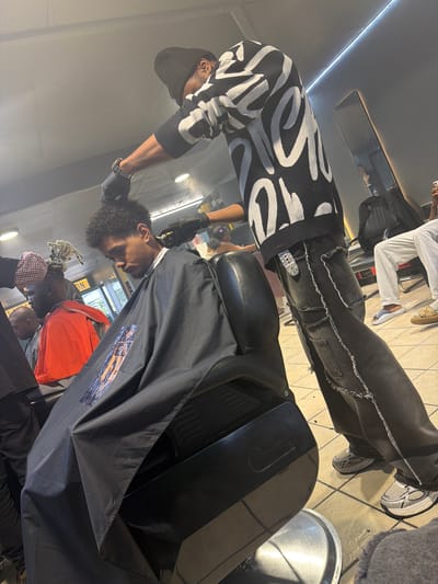 In a hair salon-like environment, a Black male, wearing a black beanie and a black and white patterned sweater, is giving a haircut to another Black male who is seated in a black salon chair and covered with a black cape. The barber is using a handheld hair clipper, clad in black gloves. Behind the barber are other people, one wearing white sweatpants and light-colored sneakers. The barber is also wearing ripped black pants with a studded belt and light-colored sneakers. Other seated people are visible in the background. The location is likely in Blouberg, South Africa, due to the prompt. The light inside is bright.