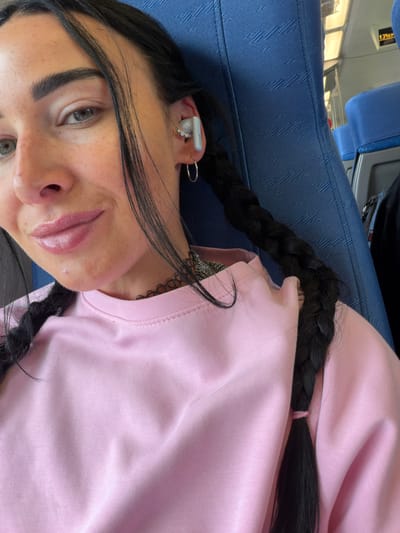This media file is a close-up, portrait-style selfie of a young person with dark hair, wearing a pink sweatshirt and a black choker necklace. The person has light-colored eyes, thick eyebrows, and appears to be wearing lip gloss. They have a white earbud in their right ear and a hoop earring in their left. One braid of their dark hair is visible, resting on their shoulder. The background suggests an indoor setting with blue seating, possibly on a train or bus, given the blue fabric and a glimpse of a window with reflections. A sign with Cyrillic text is visible in the upper left corner of the image. The image is oriented sideways.