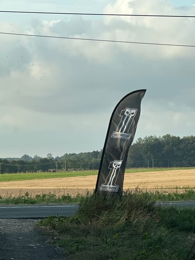 The image displays a black, feather-shaped flag with the Harley-Davidson logo printed in white. The logo features the Roman numeral "II" with a skull graphic at the top and the text "HARLEY-DAVIDSON" in a rectangular box below it. The flag is positioned in the foreground, leaning against a grassy embankment next to a road. In the background, there are fields of crops, trees, and a cloudy sky with power lines overhead. The scene appears to be an outdoor setting, possibly along a rural road. While the image is identified as being in Tôtes, France, no specific landmarks or identifying features of the city are visible in the photograph.