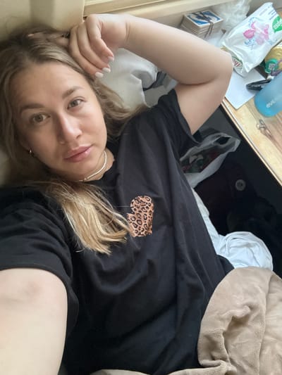 A young woman is depicted in a casual, head-and-shoulders selfie taken from a low angle while she is lying down indoors. She has light brown/blonde hair, light eyes, and fair skin, with a relaxed, slightly smiling expression. Her left arm is bent, with her hand supporting her head, showcasing light-colored nail polish. She is wearing a loose black t-shirt with a small leopard print design on the chest and a delicate silver necklace.
The setting appears to be a domestic space, likely a bedroom or living area. To her upper left, on a wooden surface, a collection of items is visible. These include a stack of small cards or tiles, bound together, with a prominent 'G' letter on one. Beside these are various personal items: a white plastic package featuring a pink flower graphic and partially visible Cyrillic text, including "абс" and "ОРГАНА"; another smaller yellow-green item; and a translucent blue water bottle with "eve" printed on it. A beige fleece blanket is draped over part of the bedding on the right side of the frame, near some darker fabrics.
The overall mood is casual and intimate, capturing a moment of rest or lounging. There are no strong indications of the time of day, but it appears to be well-lit indoors. The location context is Sergach, Russia, though no specific visual cues for this are present beyond the Russian text on the package.