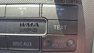 A close-up shot depicts a car radio's buttons. At the top left is a button with an arrow pointing left and the number 5. Next to it is a button with an arrow pointing right and the number 6. Below those are buttons labeled "WMA MP3" and "TEXT." The bottom row shows buttons labeled "DISC•AUX" and "TYPE FOLDER" with up and down arrows. Above these buttons is part of the phrase "AUDIO CONTROL." The buttons appear to be part of a grey or silver textured panel.