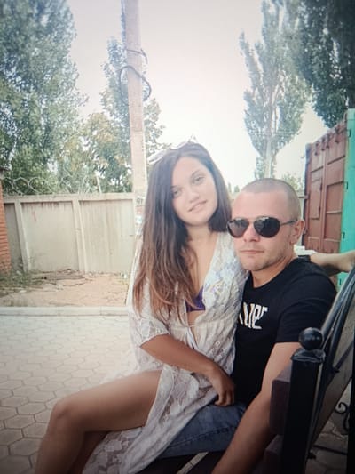 In Pishchane, Ukraine, a man and a woman are seated on a bench. The man, with a shaved head and sunglasses, wears a black t-shirt. The woman, with long brown hair, is dressed in a white lace cover-up over a purple bikini top and blue jeans. They are positioned in front of a corrugated metal fence and a concrete wall topped with barbed wire. Trees are visible in the background, and the ground is paved with hexagonal tiles. A utility pole stands to the left of the couple.