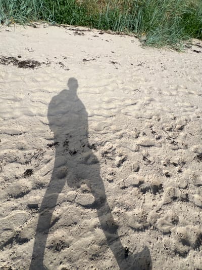 The image shows a shadow of a person on a sandy beach. The shadow is cast from the left side of the frame, and it stretches across the sand towards the right. The sand is light-colored and textured, with small ripples and indentations. There are also some dark strands of seaweed scattered on the sand. In the background, there is a strip of green grass and vegetation, likely dunes. The lighting suggests it is a sunny day. The scene appears to be a natural beach environment, not specifically within the city of Tullycullion, Ireland, as the provided context does not offer any indication of this location.