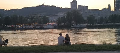 The image shows a couple sitting on the grassy bank of the Danube River in Linz, Austria, at sunset. The river's surface reflects the warm colors of the sky. In the background, across the river, is the cityscape of Linz. Visible elements include a hill with a prominent church or castle-like structure on its summit, various buildings of different architectural styles including apartment blocks and what appears to be office buildings, trees lining the riverbank, and several boats, one of which is a passenger boat with lights on. The couple is viewed from behind, with the man's arm around the woman. The scene is tranquil and romantic, capturing a moment of shared peace and contemplation of the cityscape as day transitions to evening.