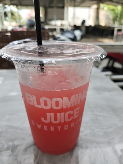In Keudealue Ie Puteh, Indonesia, a red, transparent plastic cup filled with a pink beverage and ice is positioned on a marble-patterned table. The cup is sealed with a clear plastic lid, and a black straw extends from it. White lettering on the cup reads "BLOOMING JUICE SWEET DRINK." The background is blurred but suggests an outdoor or semi-outdoor seating area with tables and chairs, possibly a cafe or restaurant. Sunlight filters through an overhead structure, creating a bright, diffused lighting effect.