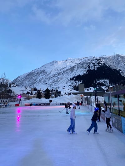 People ice skate on a rink with mountains in the background. The rink has a slight pink and blue hue, likely from the lights on the sides. One man holds a hockey stick. Other skaters are nearby, including a person wearing a blue helmet. The mountains are snow-covered and partially forested. A few buildings sit near the rink and at the base of the mountain. A bright blue sky is above.