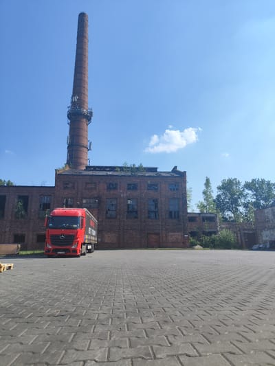 The image captures a sunny day in Częstochowa, Poland, with a prominent industrial scene. A tall, brick smokestack, weathered and topped with antennas, dominates the skyline. Adjacent to it is a large, old brick factory building, its windows broken and overgrown with vegetation, suggesting abandonment.
In the foreground, a bright red Mercedes-Benz truck is parked on a paved lot. The truck has a long, dark trailer with visible text, though it is partially obscured and difficult to read clearly. The license plate of the truck is visible, reading "JSJ 3568A".
The paved lot is made of grey interlocking bricks. To the left of the truck, a wooden pallet is visible. The sky is clear blue with a few scattered white clouds, indicating pleasant weather. The overall impression is one of a historical industrial site being utilized by modern logistics. There are no people visible in the image.