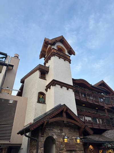A tall, beige tower with intricate wooden trim rises against a cloudy sky. The tower is part of a larger building with multi-story balconies adorned with greenery. Architectural styles vary, with some areas featuring stone facades and arched entryways. Signage reading "The Residences at Belltower" is visible on a stone wall. Below, other buildings peek out, including a round structure with a railing.