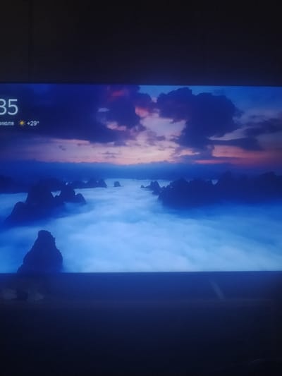 The image shows a scenic landscape with dramatic rock formations emerging from a thick layer of fog or clouds. The sky above is a gradient of deep blue, transitioning to lighter blues and purples near the horizon, with dark, textured clouds. In the upper left corner, there is text that reads "35" and "июля" followed by a sun icon and "+29°". This likely indicates the time and temperature, with "июля" being the Russian word for "July". The landscape evokes a sense of mystery and grandeur. The location is not specified within the image itself, and therefore cannot be determined to be Pshekhskaya, Russia.