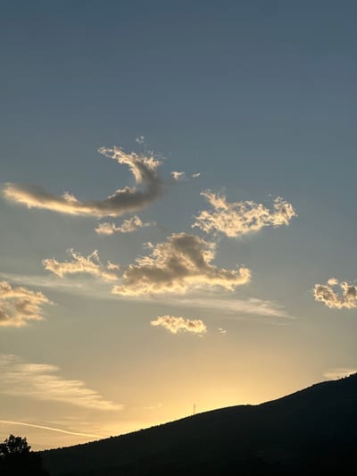 The image depicts a landscape with a sky filled with cumulus clouds. The sky transitions from a blue on the left to a golden hue on the right, likely indicating either sunrise or sunset. The right side of the image features a dark, mountainous silhouette. The photograph is rotated and would need to be turned to the right to be oriented correctly. While the city of Kemalpaşa is in Turkey, there is no evidence to establish that the location is Kemalpaşa. Without further evidence, the location cannot be determined, so the city will not be included in the description.