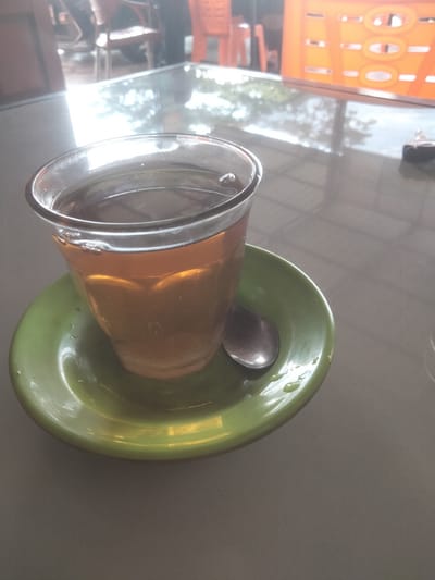The image displays a close-up of a glass of tea, likely a dark brew, resting on a glossy green saucer. A small silver spoon lies on the saucer, which also has a few visible liquid droplets. Small bubbles are present on the surface of the tea, suggesting it might be warm or freshly poured.

The setting appears to be a cafe or restaurant, indicated by the reflective table surface and blurred background elements. The table reflects bright ambient light and indistinct shapes, possibly outdoor scenery or ceiling structures. In the background, out of focus, are chairs – one brown on the left and a bright orange one on the right, alongside a distinctive orange structure or wall with circular cutout designs.

The scene is well-lit, suggesting it is daytime. No people are visible, and the image captures a static moment of a beverage ready for consumption. There is no visible text. The overall atmosphere is casual and relaxed, typical of a dining establishment in Kruengmanyang, Indonesia.