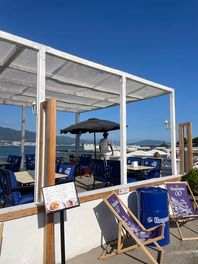 The media file depicts an outdoor seating area, likely a beach bar or restaurant, with a view of the sea and a distant coastline. The scene is bathed in bright sunlight, suggesting a clear and warm day. The architecture features a white wooden frame supporting a translucent grey canopy, providing shade.

Several deck chairs with a blue and white floral pattern, branded with "Hoegaarden 0.0", are arranged around the seating area. A large blue barrel, also branded with "Hoegaarden", is placed prominently between some chairs. Tables with chairs covered in blue cushions are visible, some set with white tablecloths. A black umbrella is positioned over a table.

In the background, the sea stretches towards a hazy coastline with mountains. A pier or walkway with structures and boats is also visible, along with a few people strolling or relaxing on the waterfront. The overall atmosphere appears casual and relaxed, typical of a seaside leisure spot.

A menu board is displayed on a stand near the seating area, showing pictures of food items, including what appears to be a dish of pasta or noodles and a plate with salmon and tomatoes. Text on the menu is in a foreign language, likely Russian. Another sign with Russian text and a prohibition symbol is visible on the white wall. A person wearing a white t-shirt with Cyrillic writing is partially visible in the middle ground, seemingly attending to the area. The presence of a few distant figures suggests the location is open for business and has patrons.