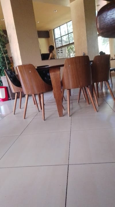 A low-angle shot shows a table with four modern chairs, three with wooden backs and one with a black cushion. A person sits at a desk visible through a window. The setting appears to be a lobby or common area with neutral-colored pillars. White tiled flooring extends to the foreground. The composition focuses on the chairs and table, creating a sense of depth.